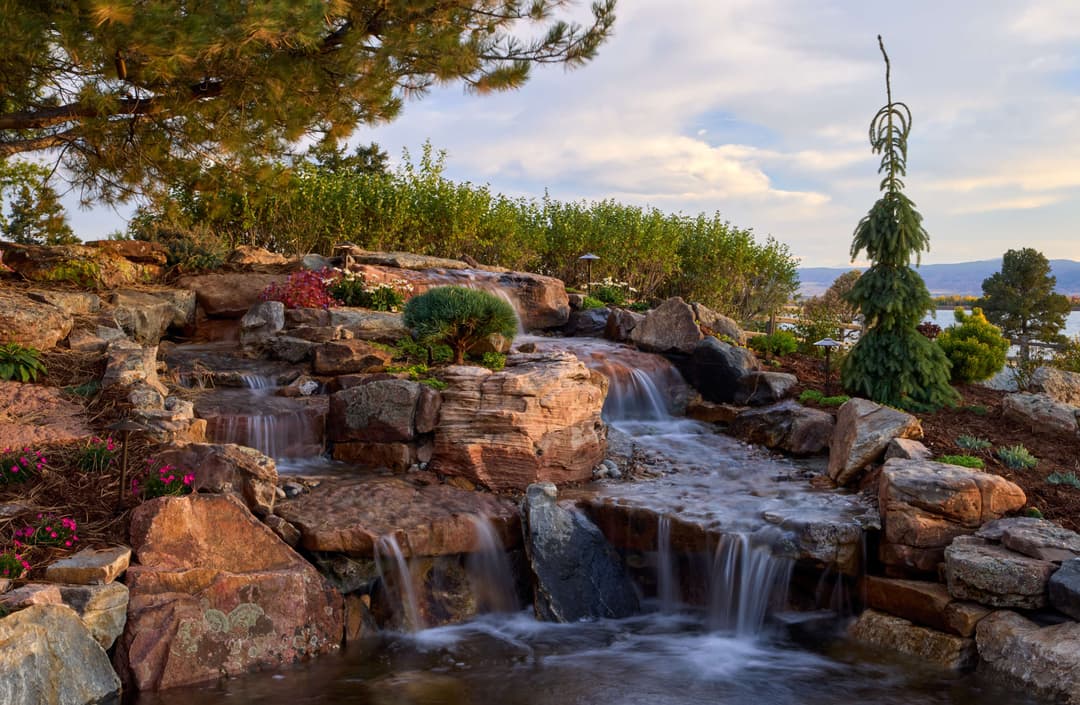 Serene waterfall cascades over rocks in a lush garden with vibrant flowers and greenery.