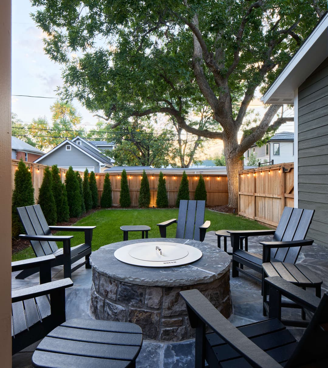 Cozy backyard fire pit with seating and string lights surrounded by greenery.