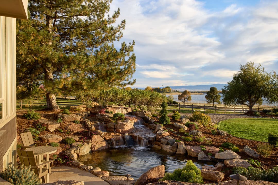 Scenic garden view with a pond, rock formations, and lush greenery by a lake at sunset.