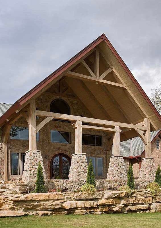 Rustic stone house with wooden beams, large windows, and landscaped garden under a cloudy sky.