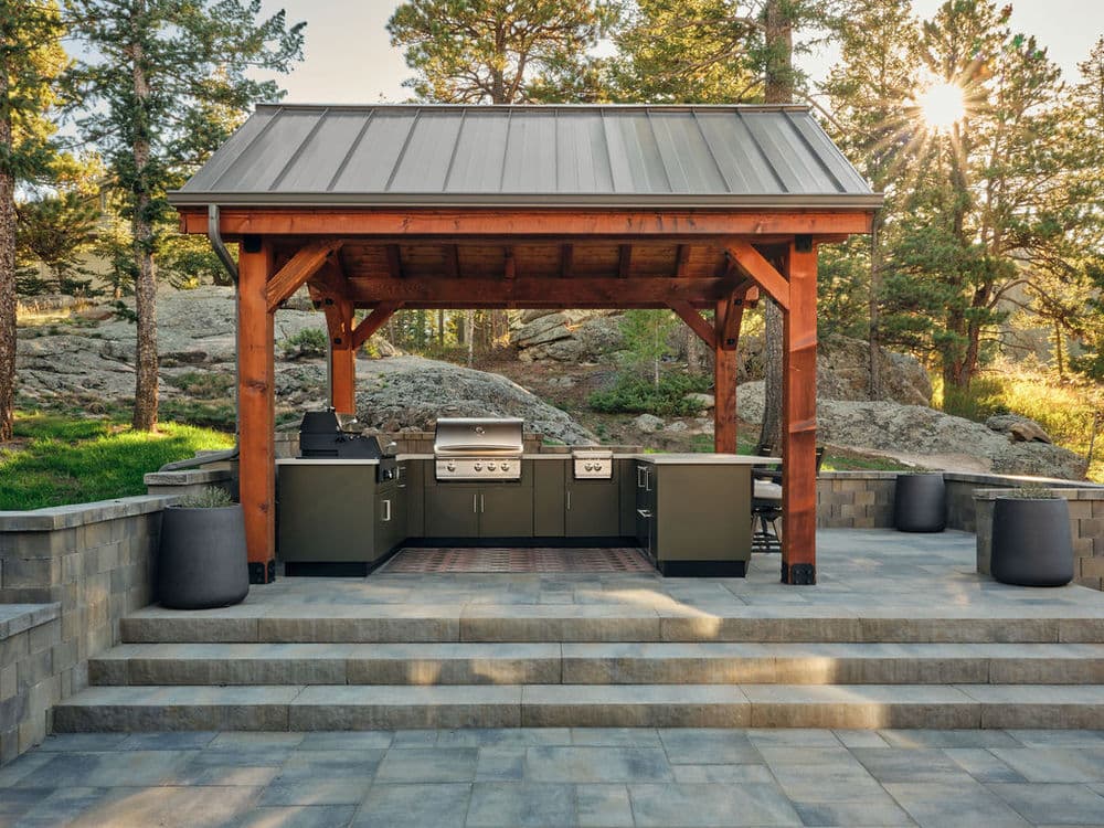 Outdoor kitchen under a wooden pavilion with modern grill and stone pathway in a serene landscape.