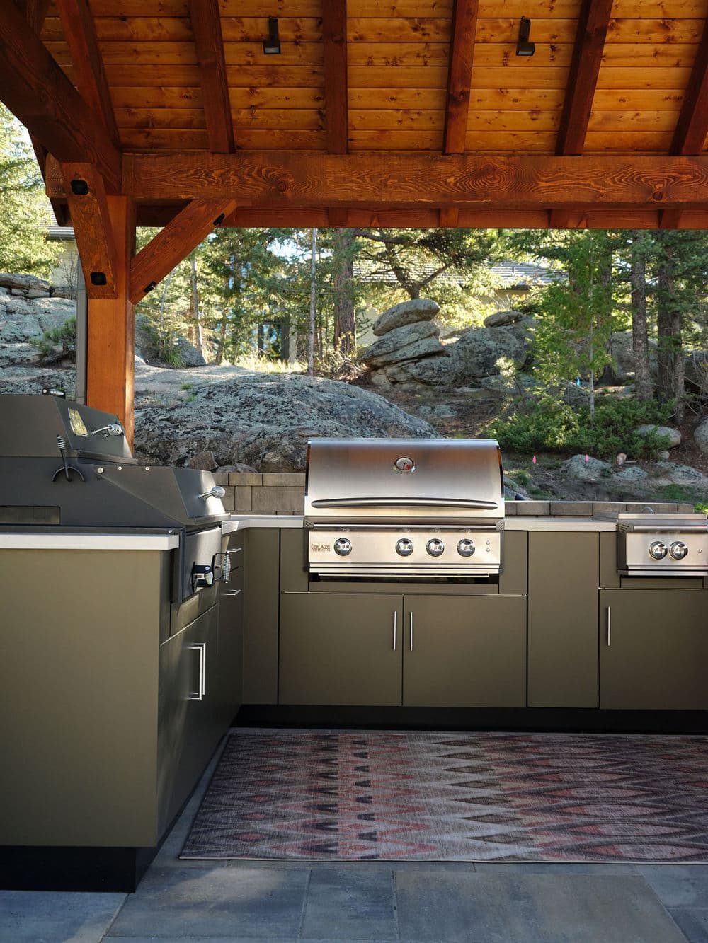 Modern outdoor kitchen with built-in grills under wooden pavilion surrounded by lush greenery.
